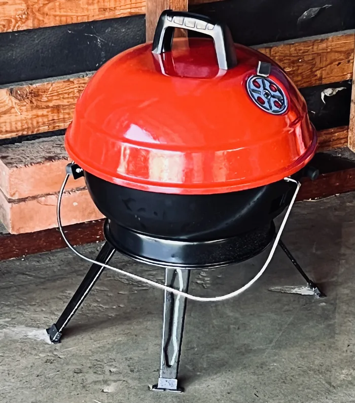 Small red portable charcoal BBQ with a domed lid and folding legs, stored in the garage