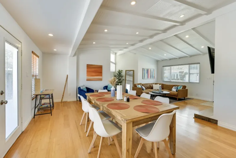 Long wooden dining table with eight white chairs in the open great room, with the lounge area beyond