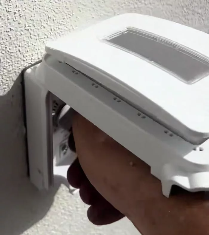 A hand reaching under the propped-open white weatherproof cover on the patio wall to turn the white igniter knob inside