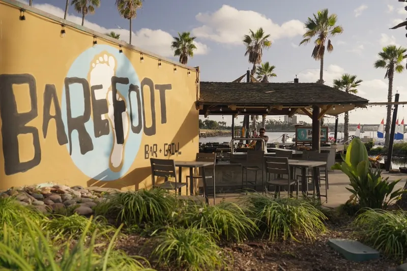 The painted Barefoot Bar & Grill sign beside the open-air bar, palm trees and Mission Bay behind it