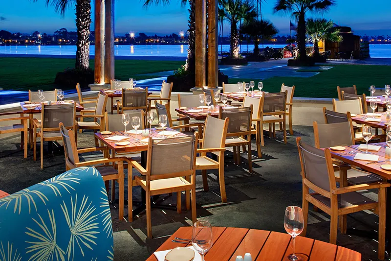 Tables set on the Catamaran’s bayfront patio at dusk, palm trees lit and Mission Bay behind
