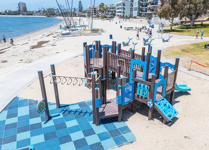 The Fanuel Street Park playground from the path, with the soft blue play surface, swings and sailboats out on the bay