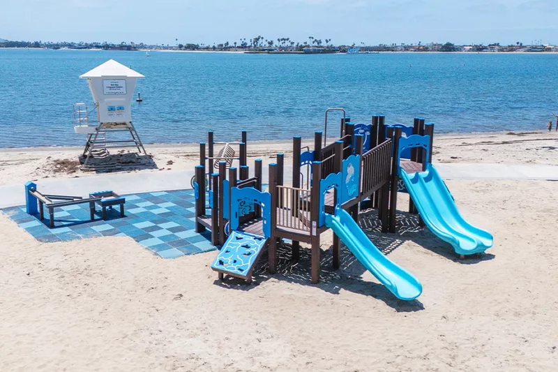 The playground at Fanuel Street Park, blue slides and a climbing frame on the sand beside the calm water of Mission Bay