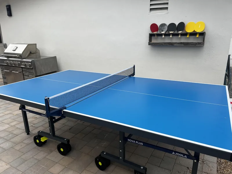 Blue ping pong table set up in the garage with paddles hanging on the wall behind